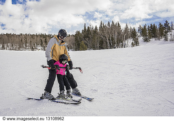 Father with daughter skiing on snow covered landscape against cloudy sky at forest