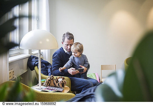 Father with smart phone while son looking at stuffed toy in hospital