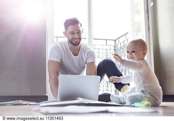 Father working on laptop and watching baby daughter playing on floor