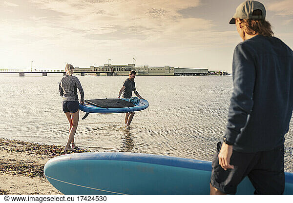 Female and male friends carrying paddleboard in sea at beach