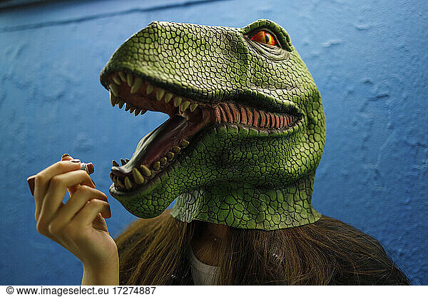 Female applying lipstick while wearing dinosaur mask against blue wall