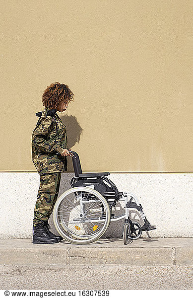 Female army soldier looking at empty wheelchair on footpath against wall