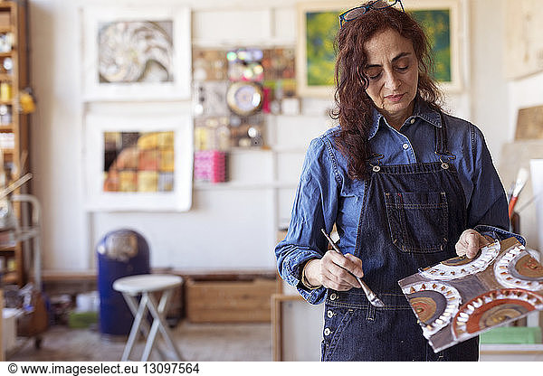 Female artist holding artwork with paintbrush in workshop
