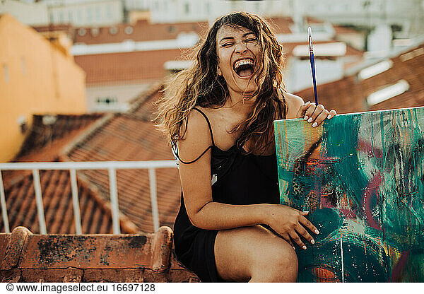 Female artist screaming while sitting on roof and holding canvas