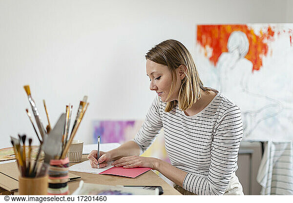 Female artist writing in book while sitting at table in home studio