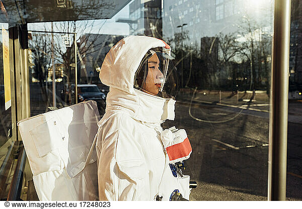 Female astronaut in space suit standing at bus stop during sunny day seen through glass