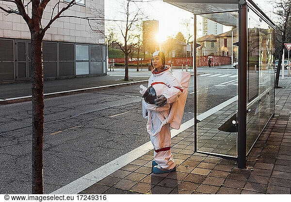 Female astronaut with space helmet standing at bus stop during sunset