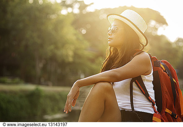 Female backpacker wearing sun hat while relaxing at riverbank on sunny day