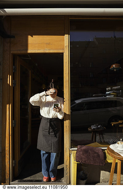 Female barista shielding eyes while using phone at doorway on sunny day