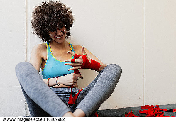 Female boxer with afro hair wrapping red bandage on hand while sitting in health club
