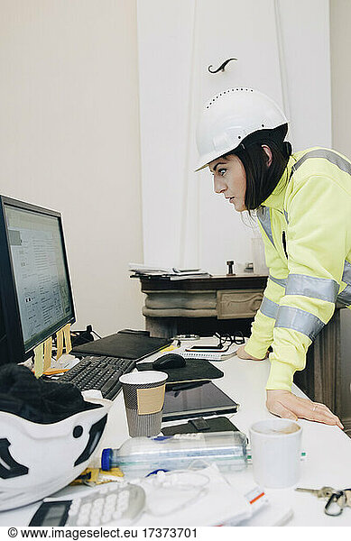Female building contractor in hardhat using computer while leaning on desk at office