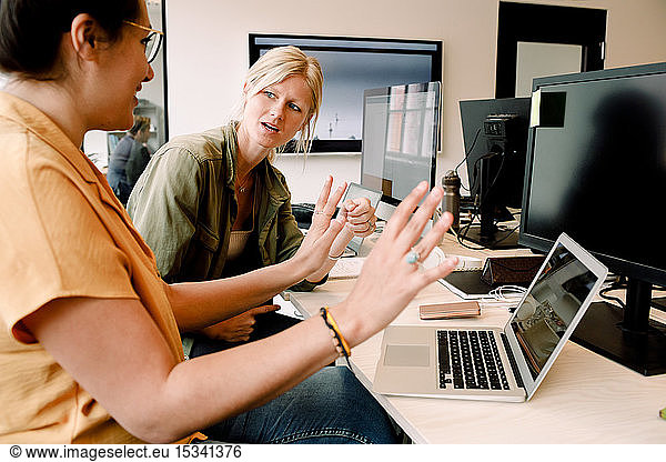 Female business professionals discussing over laptop while sitting at desk in office