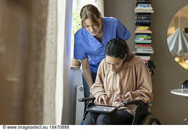 Female caregiver assisting woman using tablet PC while sitting on wheelchair at home