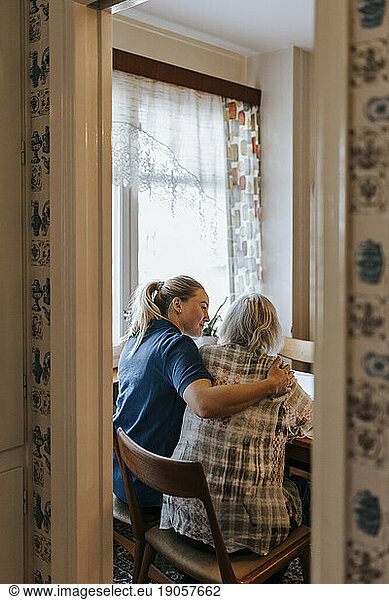 Female caregiver with arm around of senior woman sitting on chair at home