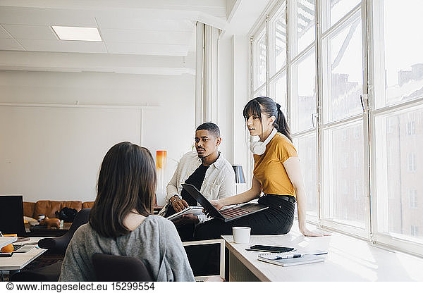 Female computer engineer discussing with colleagues while sitting on desk in office