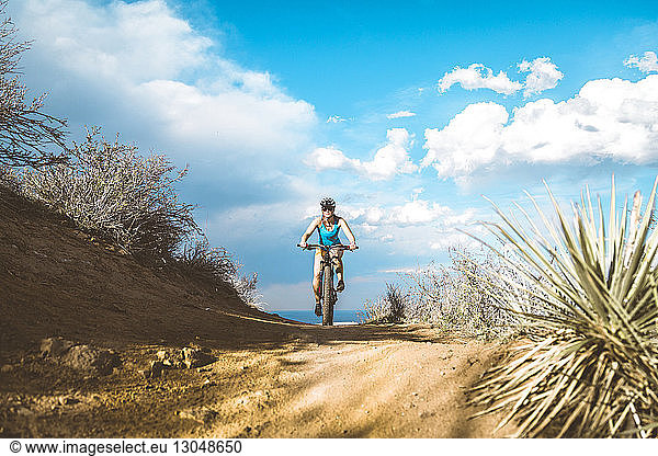 Female cyclist riding mountain bike on dirt trail against cloudy sky