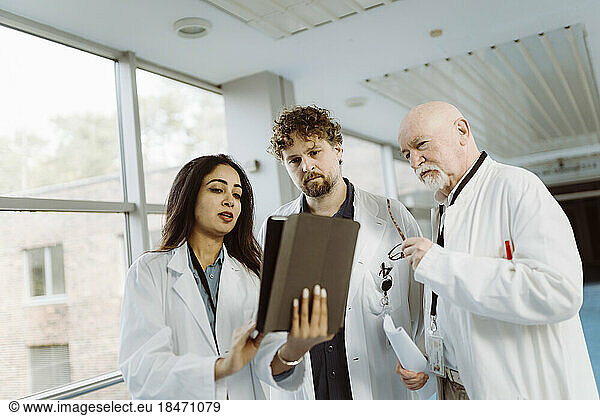 Female doctor discussing over digital tablet with male colleagues in hospital corridor