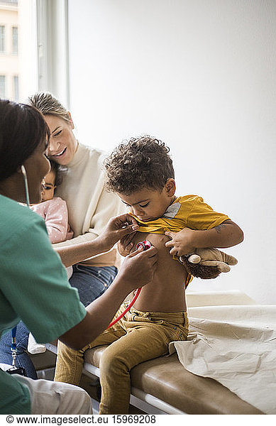 Female doctor examining boy's heartbeat with stethoscope while mother sitting and daughter in clinic