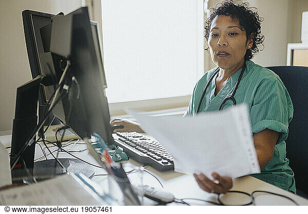 Female doctor examining medical record while using computer at desk in office