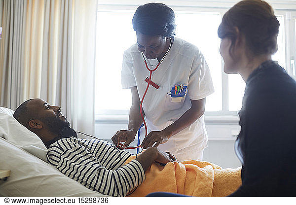 Female doctor examining patient by visitor during routine check up in hospital ward