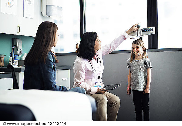 Female doctor measuring girl's height while mother sitting at clinic