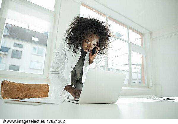 Female doctor talking on mobile phone using laptop at desk