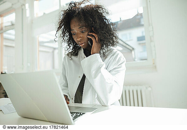 Female doctor talking on smart phone using laptop at desk