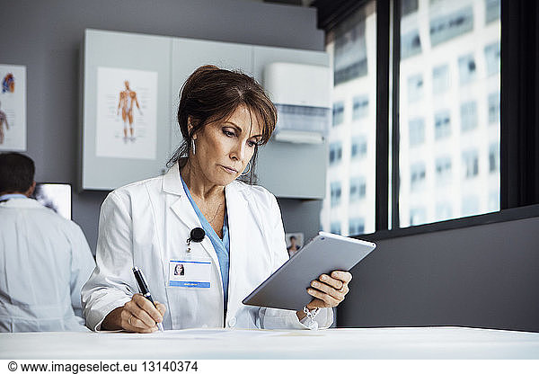 Female doctor using tablet computer while writing at desk in clinic