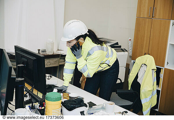Female engineer wearing hardhat talking on smart phone while leaning on desk in office
