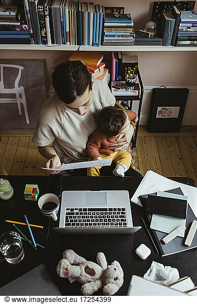 Female entrepreneur reading document while sitting with baby boy at home office