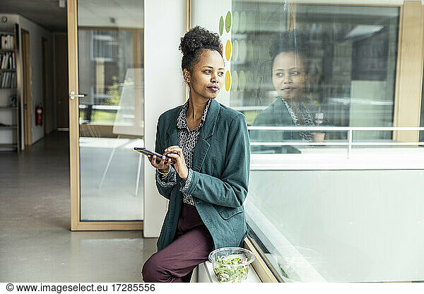 Female entrepreneur with mobile phone looking away while sitting on window sill at office