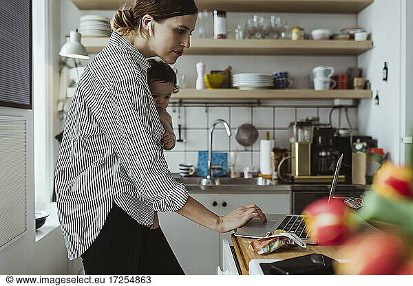 Female entrepreneur working on laptop with baby boy in kitchen