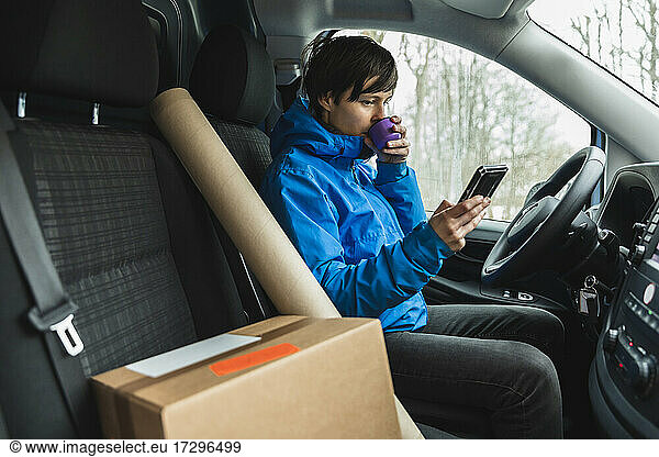 Female essential service driver drinking coffee while using smart phone in delivery van