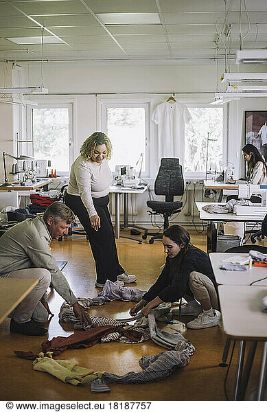Female fashion designer assisting colleagues sorting recycled clothes on floor at workshop