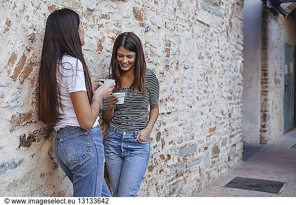 Female friends holding cappuccino talking while standing by old brick wall