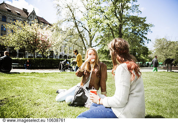 Female friends holding coffee cup while sitting on grassy field at park