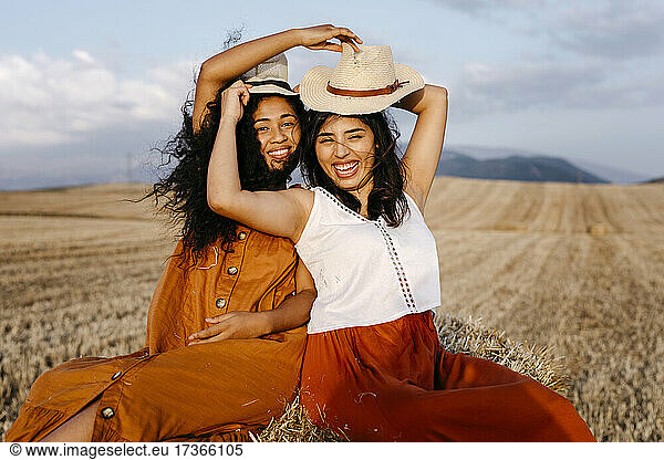 Female friends holding each other hat while sitting in farm