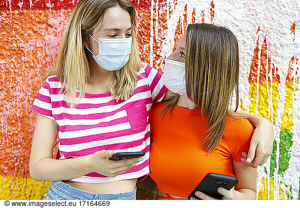 Female friends in protective face masks looking at each other while arms around against colorful wall
