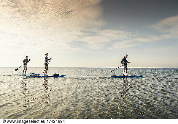 Female friends learning paddleboarding from male instructor in sea during sunset