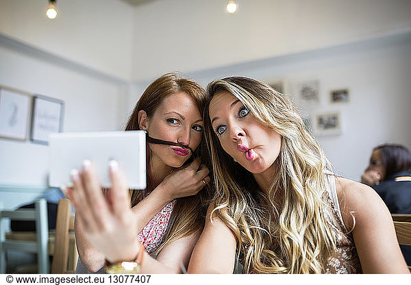 Female friends making faces while taking selfie through smart phone in cafe