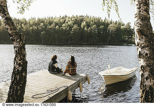 Female friends sitting on jetty over lake during sunny day
