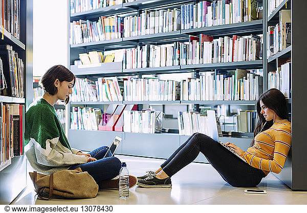 Female friends studying through laptop computers while sitting on floor in library