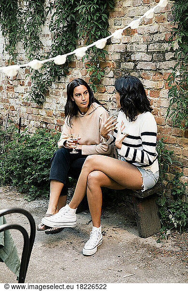 Female friends talking to each other while sitting on bench near brick wall at garden party