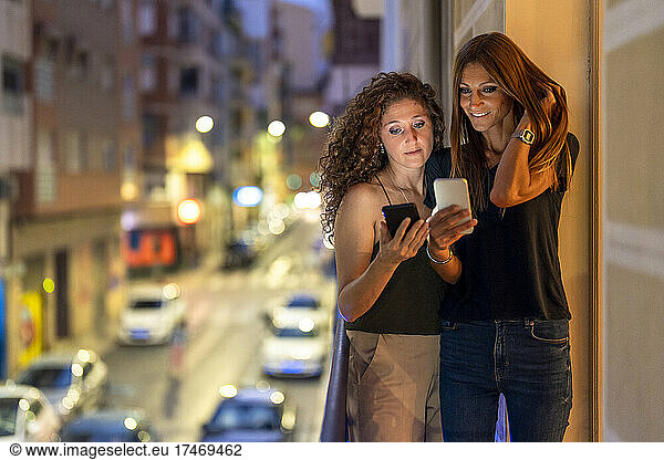 Female friends using smart phones while standing on balcony