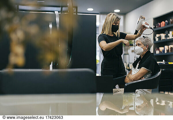 Female hairdresser with face mask blow drying customer's hair in salon