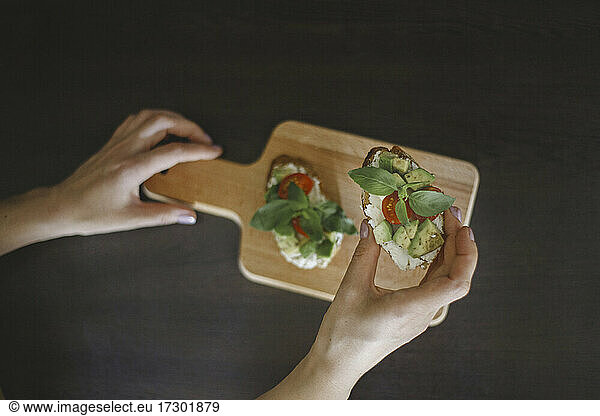 Female hand holding toast with avocado  cherry tomatoes and basil