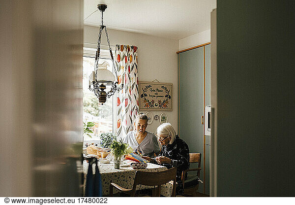 Female healthcare worker assisting senior woman to read paper in kitchen