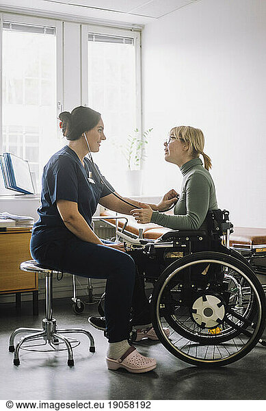 Female healthcare worker listening heartbeat of patient sitting on wheelchair in clinic