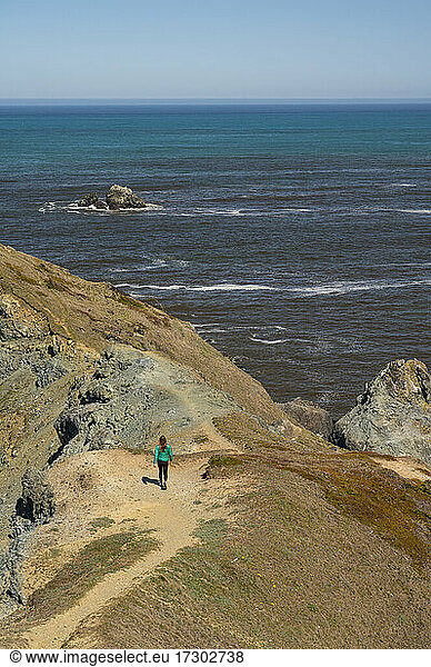 Female Hiker In Green Puffy Coat Walking Trail Leading To The Ocean