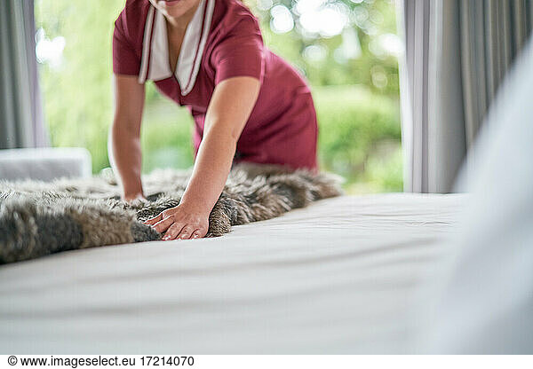 Female hotel maid arranging blanket over made bed in hotel room